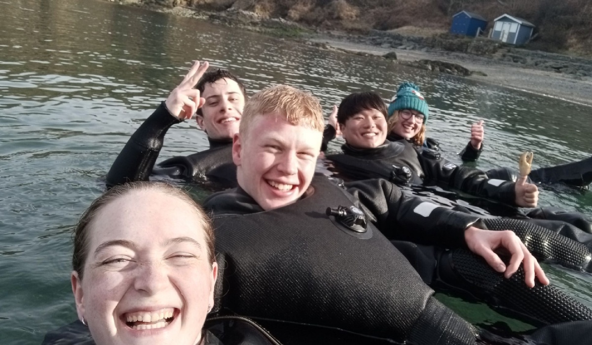 Six divers in wetsuits are standing in shallow water smiling at the camera